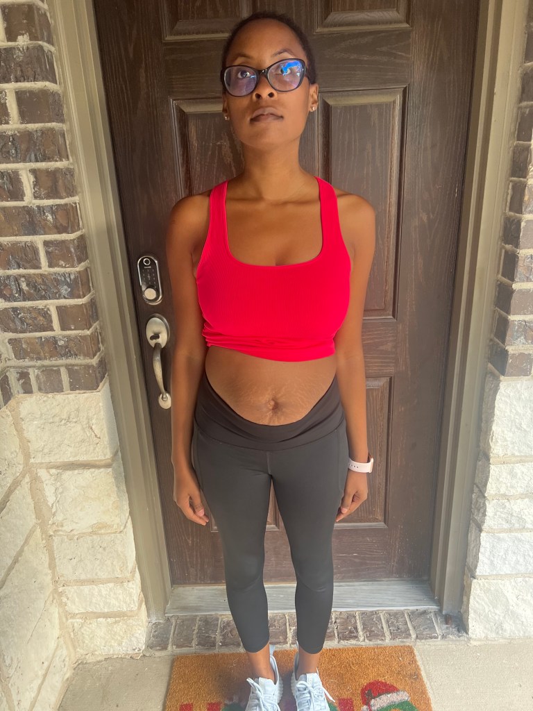 A person standing at a front door wearing a red top and black leggings, looking serious and focused. The person's top is pulled up exposing her abdomen. The background features a wooden door and brick wall.