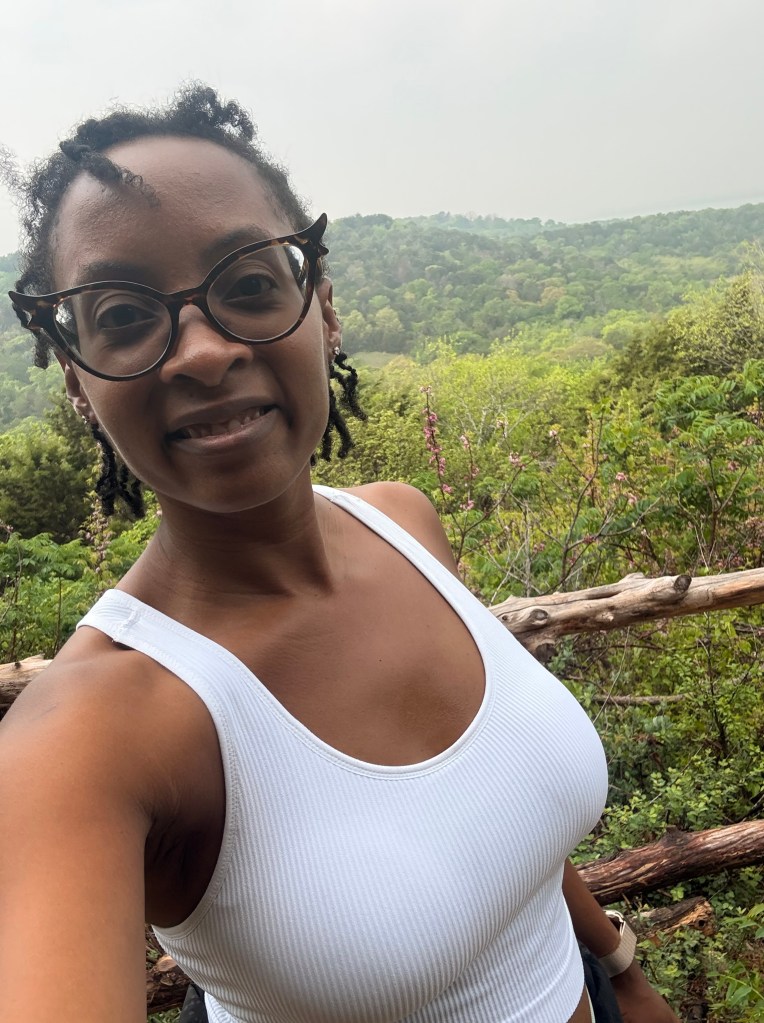 A person with glasses smiling and taking a selfie in front of a lush green landscape with trees and hills in the background.