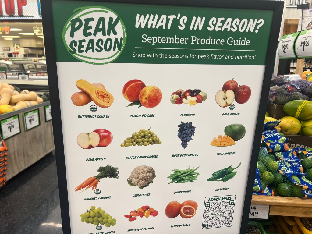 Signage displaying seasonal produce available in September, including butternut squash, yellow peaches, gala apples, and more, located in a grocery store.