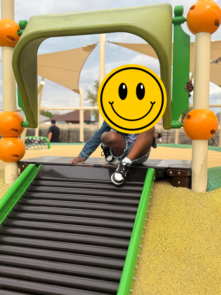 A child climbing on a playground structure with a yellow smiley face icon covering their face. The playground structure is bright, colorful and clean.