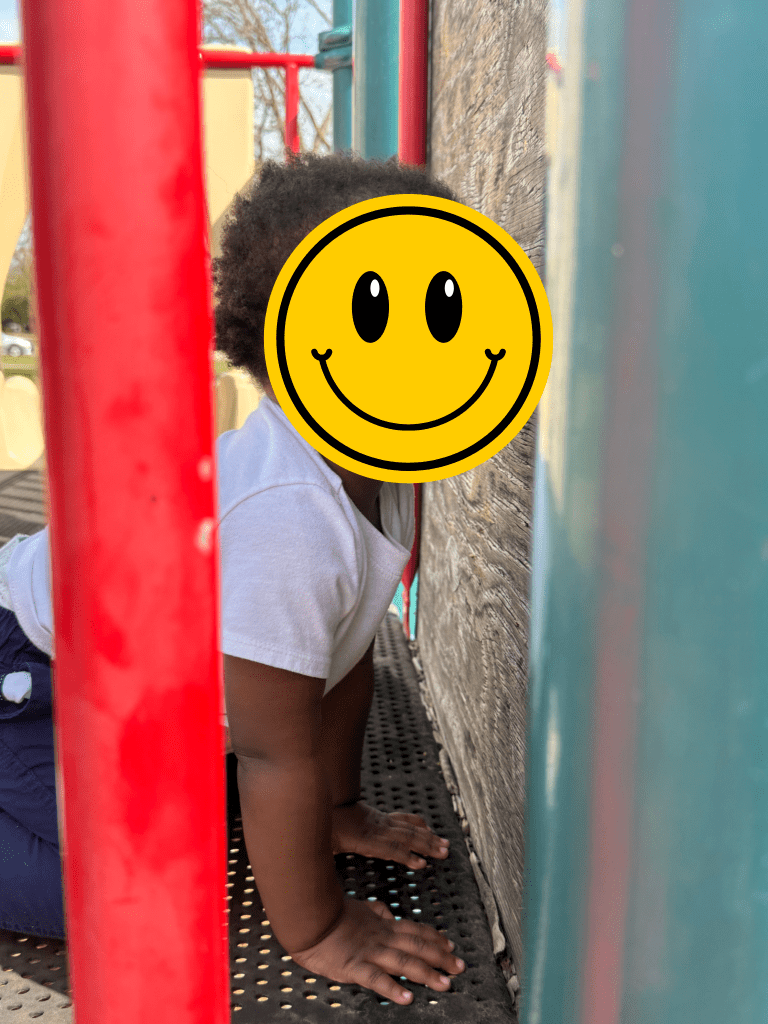 A child playing and exploring on a playground between red bars. There is a piece of wood on the play equipment replacing something that is missing. A smiling face emoji covers their identity.