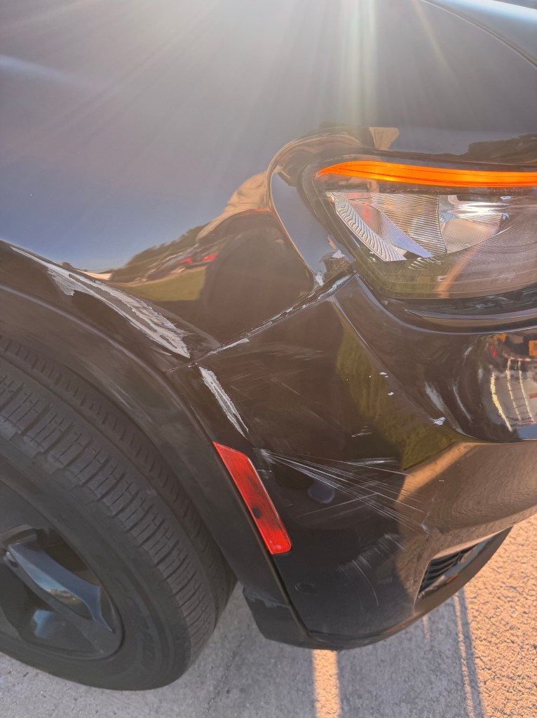 Close-up of the front passenger side of a black SUV with visible damage, including scratches and dents, and sunlight reflecting off the surface.