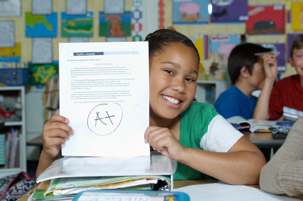 A joyful child in a classroom proudly holding up a paper with an 'A+' grade, showing her achievement while classmates are engaged in the background.