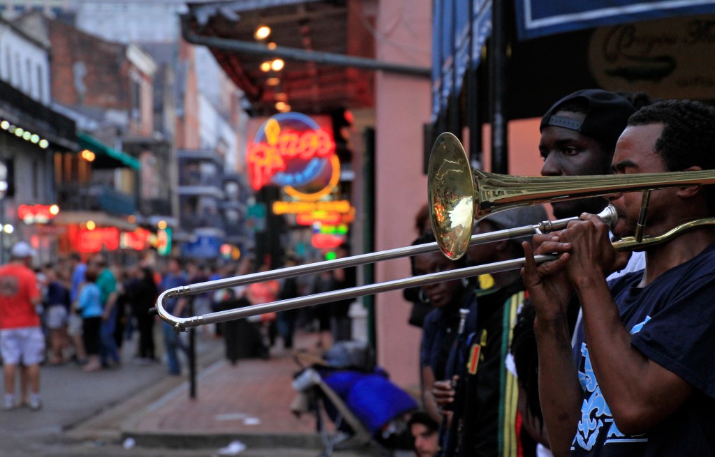 A street performer playing a trombone in New Orleans, with a lively crowd and colorful storefronts in the background.